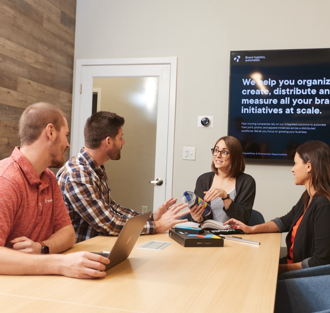 Group of people at a conference table discussing a project in front of a TV with a presentation on it.